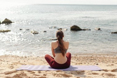 Huzurlu bir görüntü, güneşin doğuşunda kumsalda meditasyon yapan bir kadının huzurlu bir anda rahatlık ve sağlığını bulmasını gösteriyor.