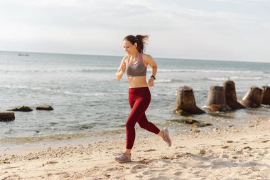 Kumsalda koşan kırmızı taytlı bir kadın. Açık gökyüzü ve okyanus dalgaları mükemmel bir açık hava aktivitesi yaratıyor..