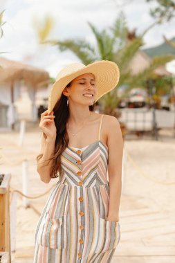 Şapkalı ve çizgili elbiseli gülümseyen kadın plajda güneşli bir günün tadını çıkarıyor.