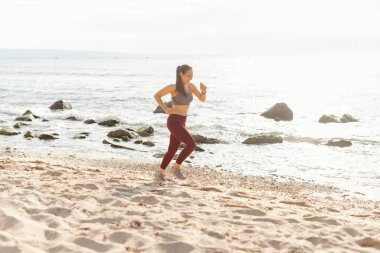 Formda bir kadın kumlu sahilde güzel bir okyanus manzarasıyla koşuyor, sağlıklı bir yaşam tarzını kucaklıyor.