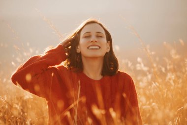 Woman in a red sweater, smiling blissfully in a golden field during sunset, capturing a moment of joy. clipart