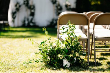 Sahnede parlak güneşin altında düzenlenen bir açık hava düğün töreninde sıra sıra sandalyeler ve çiçek süslemeleri yer alıyor.