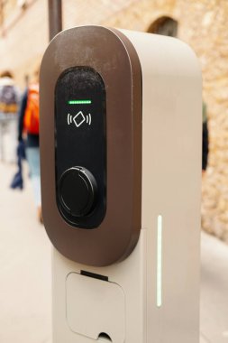 Şehrin arka planında insanların bulunduğu bir caddede modern bir elektrikli araç şarj istasyonuna yakın plan..