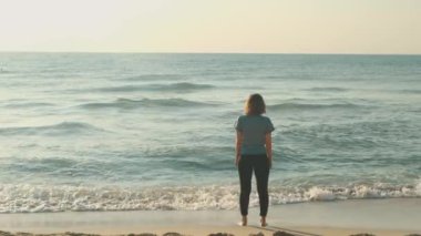 Huzurlu bir görüntü, sakin bir günbatımı sırasında kumlu bir plajda tek başına okyanus dalgalarını seyreden bir kadını gözler önüne seriyor.