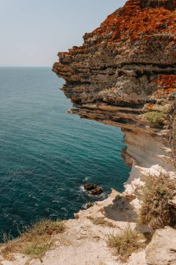 Kıyı sahnesi, kristal mavi denize bakan engebeli bir uçurumu resmediyor. Doğal güzellik ve sükuneti simgeliyor.