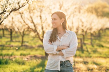 Gün batımında çiçek açan bir bahçede, beyaz bir gömlek ve kot giyen, baharın tadını çıkaran bir kadın..