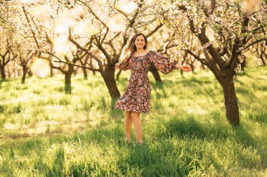 Güneşli bir bahçede, çiçekli bir elbise giymiş neşeli bir kadın baharın başlangıcını işaret ederek sevinçle dönüyor.