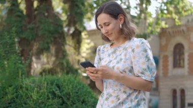 Genç bir kadın etkin bir şekilde akıllı telefonuyla meşgul. Etrafı yemyeşil, modern yaşam tarzı eğilimleriyle çevrili.