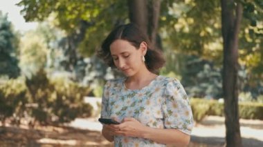 Bir kadın, hayat dolu, yemyeşil bir parkta yürürken rastgele akıllı telefonunu kullanır.