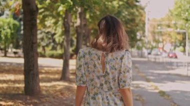 Çiçekli elbiseli bir kadın canlı renkler ve dökülen yapraklarla dolu bir sonbahar parkında geziniyor.