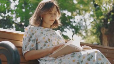 Genç bir kadın yemyeşil bir parktaki ahşap bir bankta büyüleyici bir kitap okurken hevesle saf neşe buluyor.