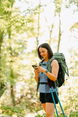 Sakin bir ormanda bir kadın akıllı telefonunu kontrol eder. Sırt çantası ve yürüyüş direğiyle maceraya hazırdır.