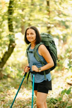 Yürüyüş direkleri ile donatılmış bir kadın şaşırtıcı derecede güzel yeşil bir ormanda yürüyüşünün tadını çıkarıyor.