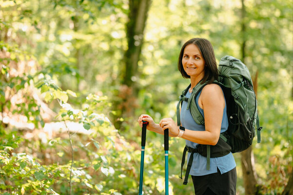 A woman hikes in a beautiful green forest, equipped with sturdy trekking poles and a heavy backpack