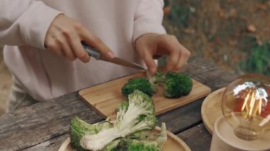 Bir insan, doğal elementlerle çevrili taşralı ahşap bir masada ustalıkla taze brokoli kesiyor.