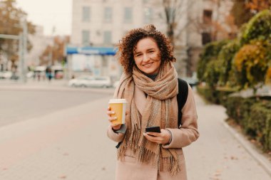 Neşeli bir kadın mutlu bir şekilde sıcak kahvesini yudumlarken, sonbahar kıyafetleri içinde, canlı bir sonbahar manzarası ile çevrili.