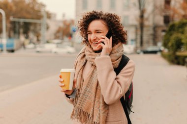 Neşeli genç bir kadın, şehrin hareketli caddesinde elinde dumanı tüten bir kahve bardağıyla telefonuyla mutlu bir şekilde konuşuyor.