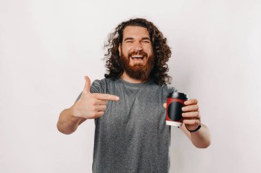 Neşeli bir adam enerjik bir şekilde dumanı tüten bir kahve fincanını işaret eder. Parlak ve canlı bir ortamda pozitiflik saçar.