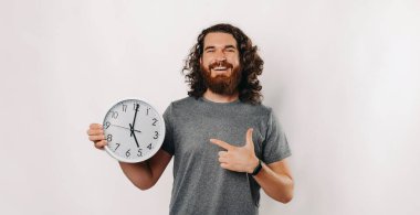 Bakımlı sakallı neşeli bir adam saati heyecanla işaret ediyor, zamanı ve pozitifliği simgeliyor.