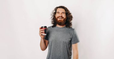 Kıvırcık saçlı, gülümseyen bir adam temiz, parlak bir arkaplanda dururken ferahlatıcı bir içkiden zevk alır.