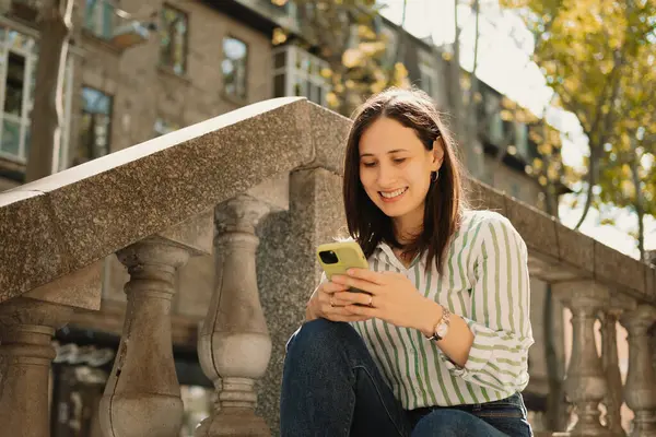 Moda kıyafetleri içinde neşeli bir kadın akıllı telefonunu güneşli, canlı bir şehir manzarasında bir taş korkuluğun üzerinde kullanır.