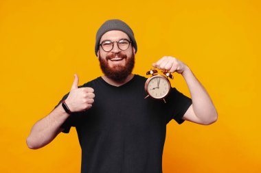 Neşeli bir adam, parlak bir çalar saati parlak sarı bir arka plana karşı tutarken başparmağını kaldırır.