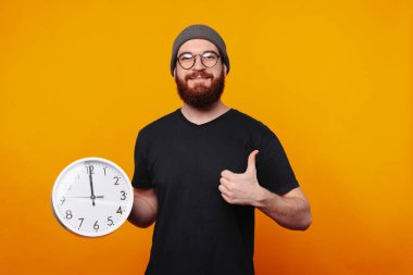 Neşeli genç bir adam, parlak sarı bir arka plana karşı saati tutarken, sevinçten havalara uçuyor.