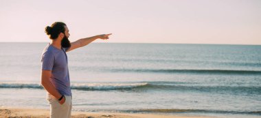 Düşünceli bir adam sakin bir sahilde ufka doğru hareket eder, derin bir yansıma anı yakalar.