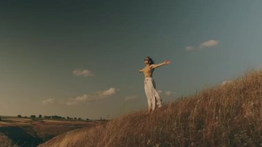 Bir kadın zarif bir şekilde bir tepede duruyor, kolları açık, etrafındaki güzel doğayı kucaklıyor.