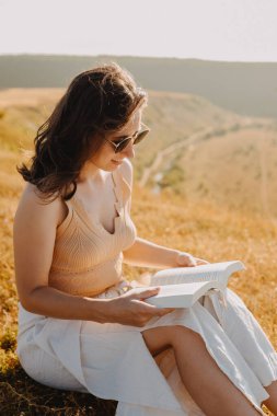 Bir kadın, sakin bir çayırın ılık güneş ışığında doğa ve barışı kucaklayan büyüleyici bir kitap okumaktan zevk alır.