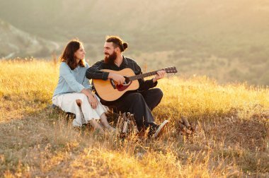 Sevgi dolu bir çift doğada güzel bir romantik anı paylaşıyor, çarpıcı bir günbatımında akustik gitar çalıyorlar.
