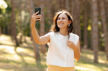 Genç bir kadın güneşli bir ormanda neşeli bir selfie çeker, neşeli ruhunu ve mutluluğunu gösterir.
