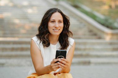 Neşeli genç bir kadın dışarıda merdivenlerde oturuyor, mutlu bir şekilde dışarıda sakin ve huzurlu bir şekilde akıllı telefonunu kullanıyor.