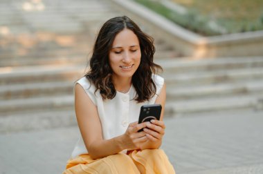 Bir kadın dışarıda merdivenlerde oturmuş, akıllı telefonuna sevinçle gülümserken doğada huzurlu bir anın tadını çıkarıyor.