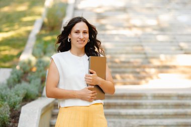 Kendinden emin ve neşeli bir kadın, elinde bir klasör tutarken neşeli bir şekilde gülümsüyor, güneşli, canlı bir günde dışarıda yürüyor.