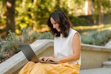 Süslü, beyaz bluzlu ve parlak sarı etekli bir kadın laptopuyla dışarıda sakin bir şekilde çalışıyor.