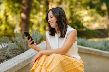 Neşeli bir kadın açık hava ortamında, yemyeşil bir ortamda akıllı telefonunun keyfini çıkartıyor.