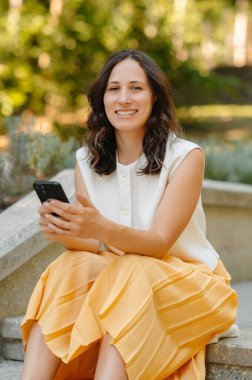 Genç bir kadın dışarıda canlı bir ortamda oturuyor, akıllı telefonuyla uğraşırken ışıl ışıl gülümsüyor.