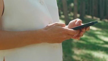 Genç bir kadın açık havada, gür ağaçlar ve canlı doğa ile çevrili iken akıllı telefonuyla aktif olarak meşgul.