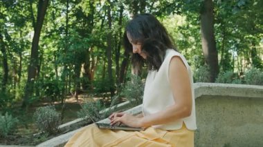 Genç bir kadın, sakin ve huzurlu bir orman ortamında otururken dizüstü bilgisayarında özenle yazı yazıyor.