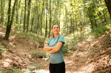Kendinden emin ve güçlü bir kadın, güneşli bir ormanda gururla duruyor. Etrafındaki doğanın güzelliğini tamamen kucaklıyor.