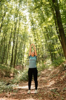 Huzurlu bir ormanda, bir kadın uzun ağaçlar ve yeşilliklerle çevrili güneşin altında zarifçe uzanır.