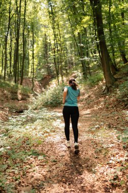Bir kadın, etrafındaki yemyeşil sularla çevrili güneşli bir orman yolunda huzurlu ve huzurlu bir yürüyüşün tadını çıkarır.