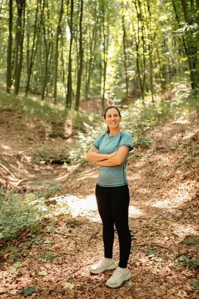 Güler yüzlü bir kadın yemyeşil bir ormanda duruyor, güneş ışığı uzun ağaçların arasından süzülüyor.