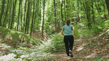 Bir kadın, etrafını çevreleyen canlı yeşil ağaçlar ve doğanın kendisi ile çevrili sakin bir orman patikasında düzenli olarak koşar.