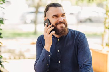 Mavi tişörtlü gülümseyen bir adam açık bir kafede telefonla konuşuyor, pozitif enerji yayıyor.