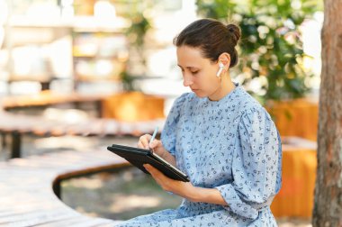 Mavi elbiseli bir kadın açık havada huzur içinde oturuyor. Etrafı yemyeşil. Fikirlerini yazıyor.