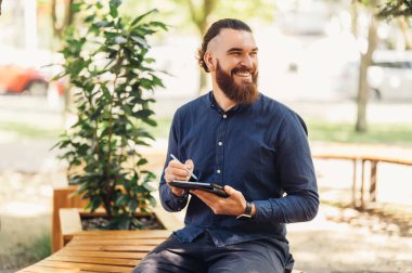 Neşeli sakallı bir adam mutlu bir şekilde parkta bir bankta oturuyor, açık hava ortamında tablet kullanıyor.