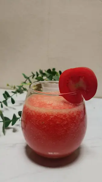 Bir bardak domates suyu ve beyaz bir masanın üstündeki bardağın dudağında domates dilimi.