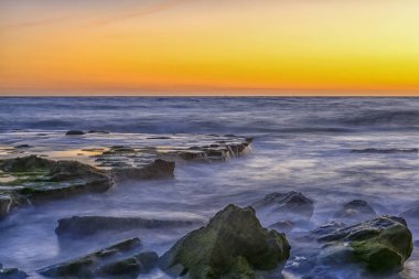 Dramatik bir günbatımı gökyüzü sisli, kayalarla kaplı bir sahile ılık renkler salıyor..